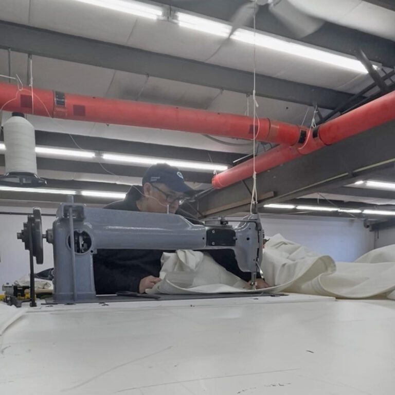 A sailmaker at Ullman Sails Northeast working on a white sail at an industrial sewing machine inside the sail loft.