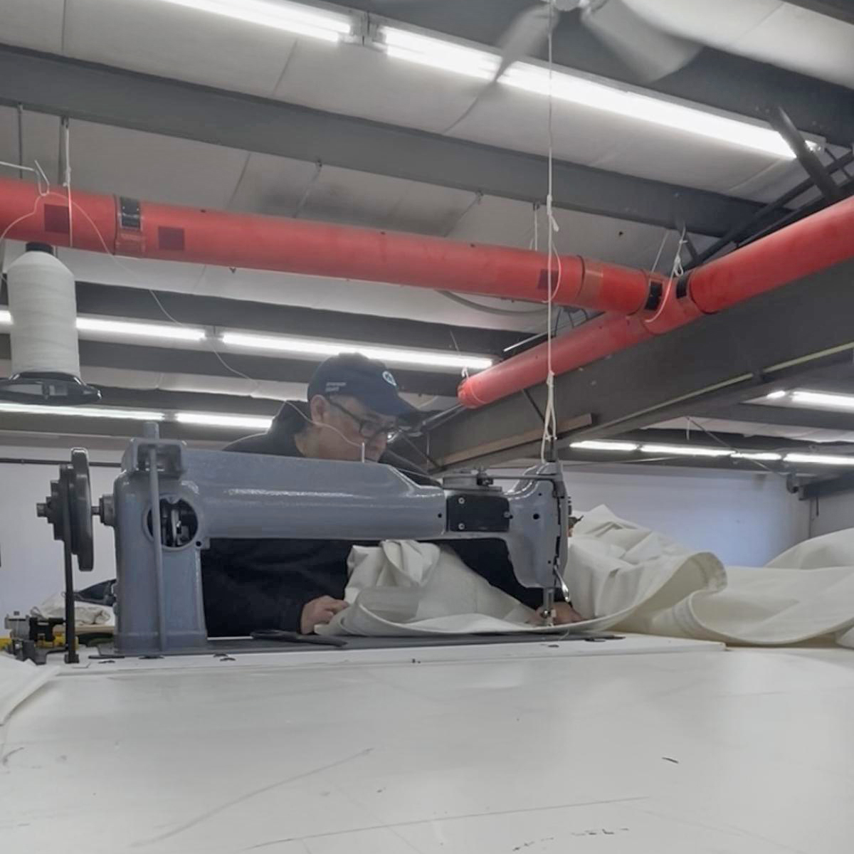 A sailmaker at Ullman Sails Northeast working on a white sail at an industrial sewing machine inside the sail loft.